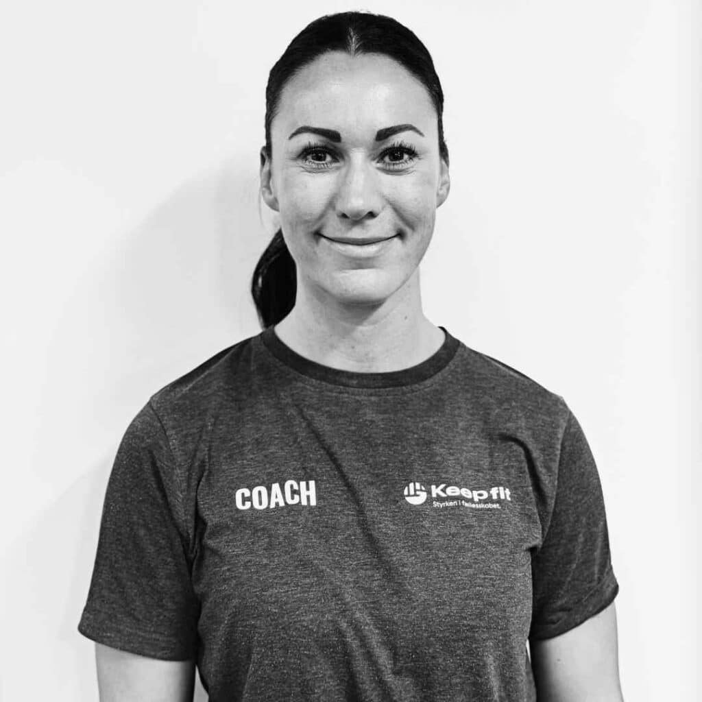 Natasja Keep står op ad en hvid væg og kigger ind i kameratet iført t-shirt med logo Keep Fit på det ene bryst og teksten Coach på det andet bryst